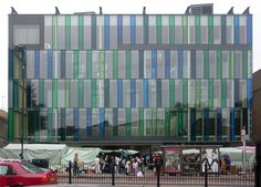 a red car is parked in front of a building with many windows and people walking around