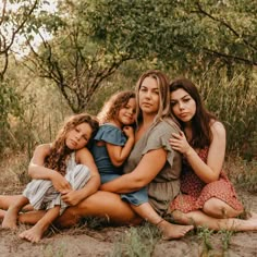 three women and two children are sitting on the ground in front of some trees with their arms around each other