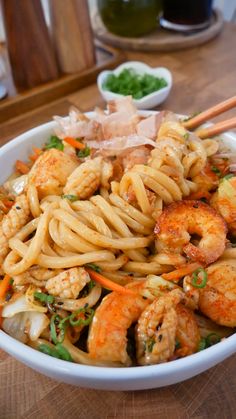 a white bowl filled with noodles and shrimp next to chopsticks on a wooden table