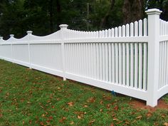 a white picket fence in the grass with leaves on the ground and trees behind it