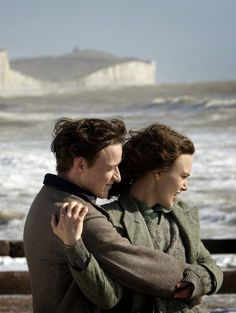 two people embracing each other near the ocean and white cliffs in the background, with waves crashing on them