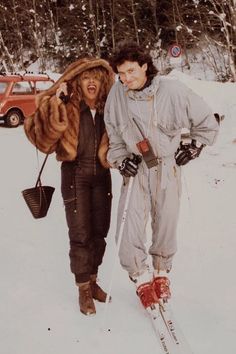 two people standing in the snow with skis on their feet and one person wearing a fur coat