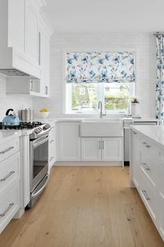 a kitchen with white cabinets and wood flooring next to a window that has blue flowers on the valance