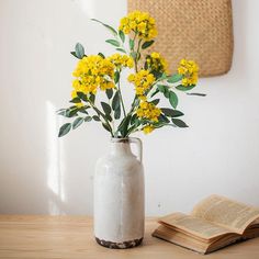 a vase with yellow flowers sitting on a table next to an open book and straw bag