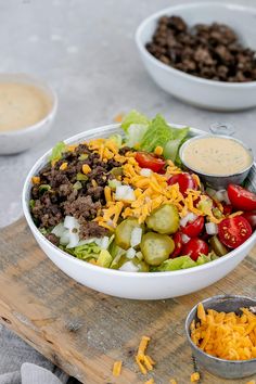 a salad with cheese, meat and vegetables on a wooden cutting board next to bowls of dressing