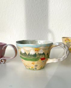 three coffee cups sitting on top of a table next to each other, one painted with farm scenes and the other decorated with flowers