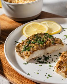 a white plate topped with fish next to a bowl of rice and lemon wedges