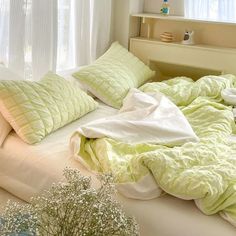 an unmade bed with green sheets and pillows on it, in front of a window