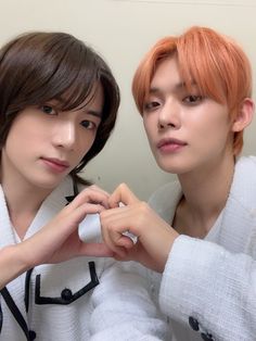 two young men sitting next to each other in front of a white wall, one with orange hair