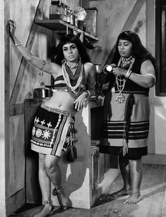 two women dressed in native american clothing standing next to an open door with pots on the stove