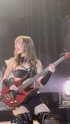 a woman in leather clothes holding a red guitar on stage with other instruments behind her