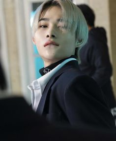 a young man with blonde hair wearing a black suit and white shirt, looking off into the distance