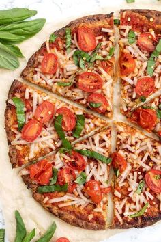 a pizza with cheese, tomatoes and basil on it sitting on a table next to green leaves