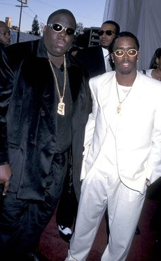 two men standing next to each other in white suits and sunglasses on the red carpet