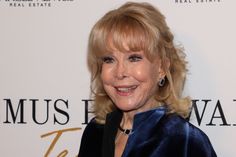 an older woman with blonde hair and blue velvet jacket smiling at the camera while standing in front of a white backdrop