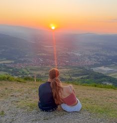 a man and woman sitting on top of a hill watching the sun go down in the distance