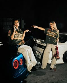 two women standing next to each other in front of a car talking on their cell phones