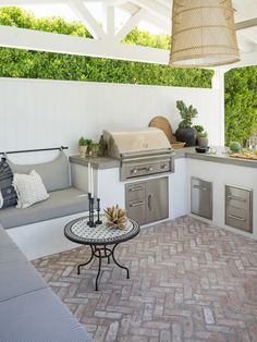 an outdoor kitchen with grill, sink and table on brick flooring next to white walls