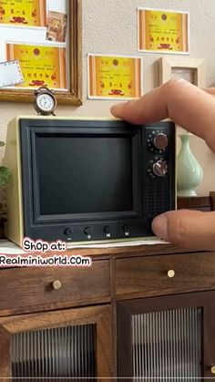 a hand is holding a small toy tv in front of a dresser with pictures on the wall behind it