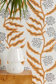 a white vase sitting on top of a wooden table next to a wall with orange and black designs