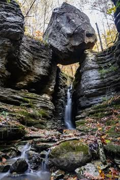 a small waterfall running between two large rocks in the woods with leaves on the ground