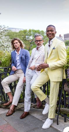 three men in suits are sitting on a bench and one man is wearing white shoes