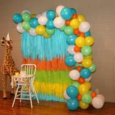 a giraffe standing in front of a balloon arch with streamers and balloons