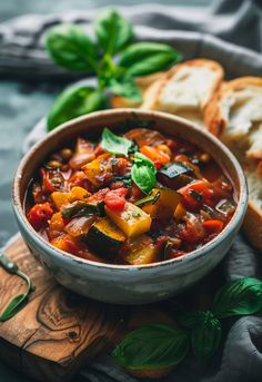 a bowl filled with vegetable stew next to bread and basil leaves on a cutting board