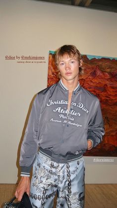 a young man standing in front of a painting holding a black bag and looking at the camera