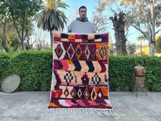 a man is holding up a multicolored rug in front of some bushes and trees