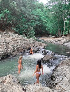 two women in bikinis are wading through the water near some rocks and trees