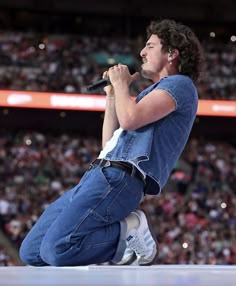 a man sitting on top of a stage holding a microphone in his right hand and wearing blue jeans