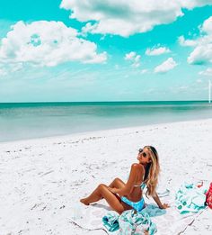 a woman sitting on top of a beach under a blue sky and white cloud filled sky