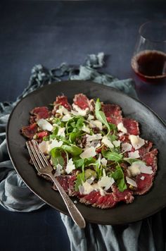 a black plate topped with meat and veggies next to a glass of wine