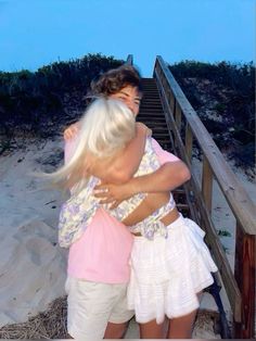 two women hugging each other while standing on a wooden walkway over sand dunes at the beach