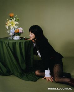 a woman sitting on the ground next to a table with a vase and flowers in it