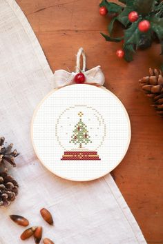 a cross - stitch ornament with a christmas tree on it next to pine cones