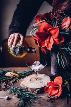 a person pouring an alcoholic drink into a wine glass on a table with flowers and greenery