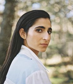 a woman with long black hair wearing a white shirt and blue jacket in the woods