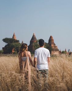 a man and woman holding hands walking through tall grass in front of pagodas on a sunny day