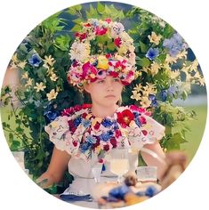 a woman with flowers on her head sitting at a table in front of some plants