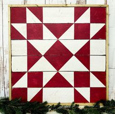 a red and white quilt is hanging on a wooden wall next to greenery in front of it