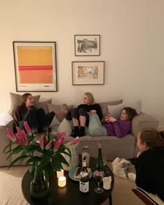 three women sitting on a couch with bottles of wine in front of them and two vases filled with flowers