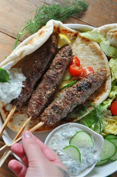 a person holding two skewers over a plate of food with cucumbers, lettuce and tomatoes