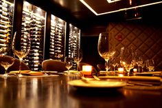 a dimly lit table with wine bottles and glasses in the background, along with place settings for dinner