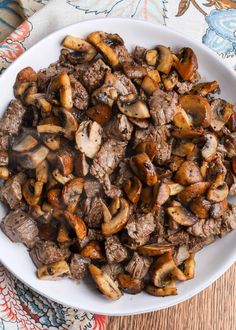 a white plate filled with meat and mushrooms on top of a wooden table next to a colorful cloth