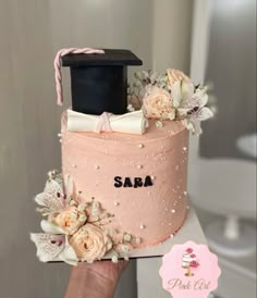 a hand holding a pink cake decorated with flowers and a graduation cap on top that says saba