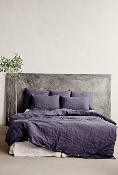 a bed with purple linens and pillows in a white room next to a potted plant