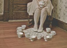 a woman is sitting on a chair with cups around her and holding a tea pot