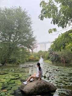 a woman sitting on top of a rock next to a pond filled with lily pads